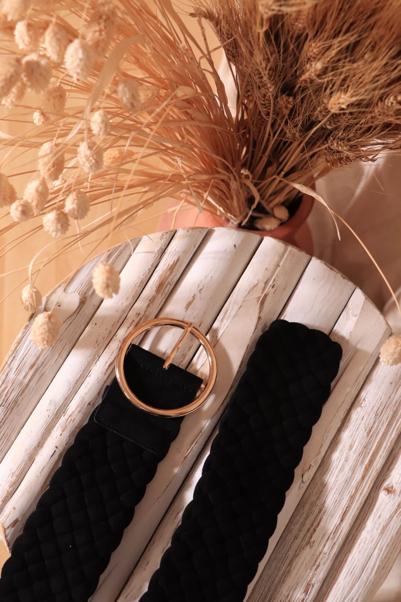 ceinture Adèle noir boucle or posé sur une table en bois blanc et vase avec feuilles