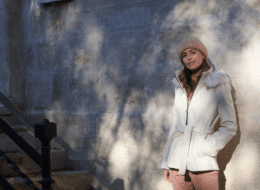 quel manteau tendance choisir cet hiver ? mannequin blonde adossée contre un mur avec un manteau Actuelle blanc longueur 3/4 et capuche fausse fourrure noué à la taille 